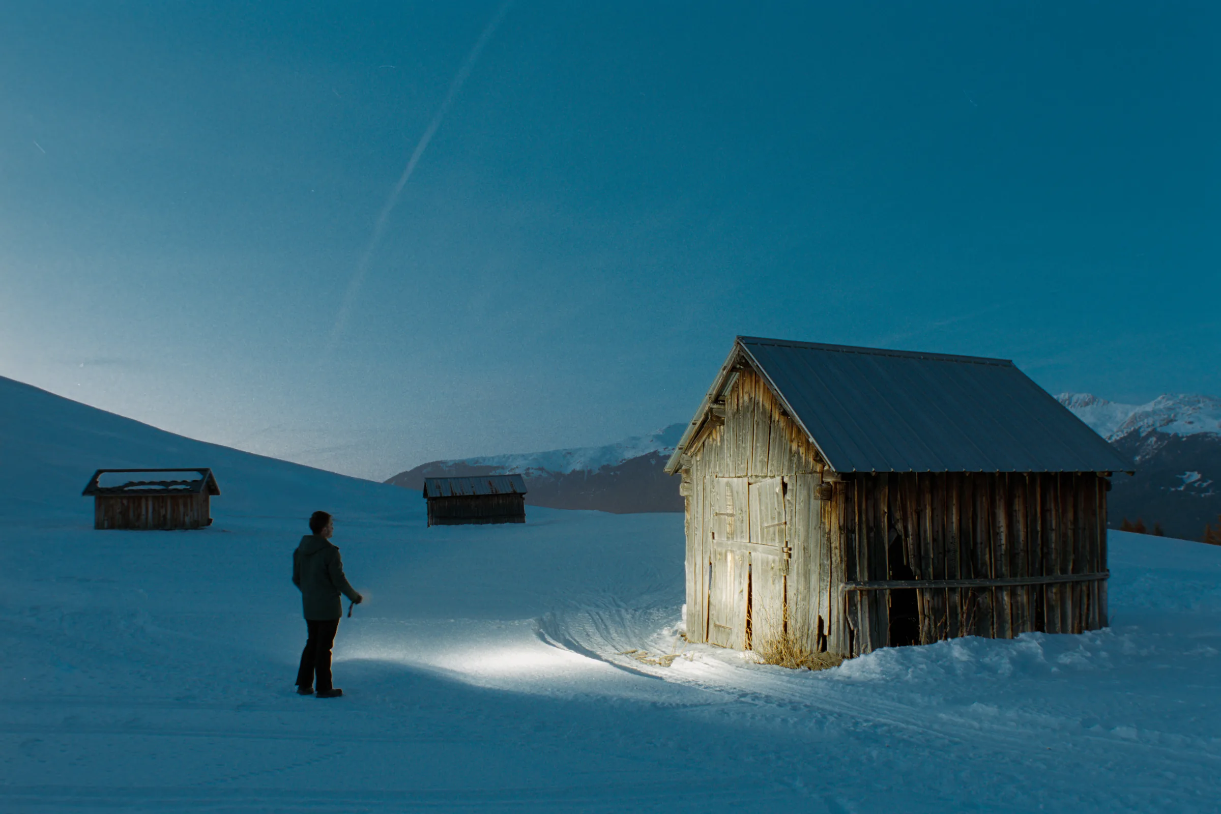 A lone figure facing a wooden barn lit on a snowy alpine night, a long-exposure planning subject